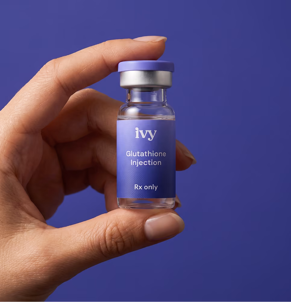 Hand holding a small clear glass vial with a purple label and cap, labeled 'ivy Glutathione Injection Rx only' against a purple background.