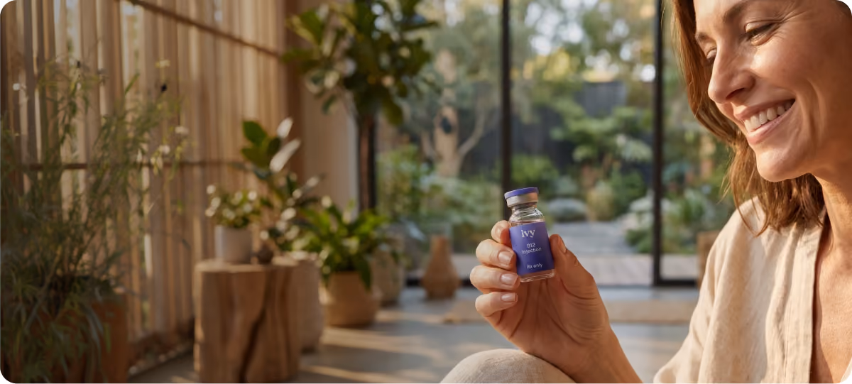 A smiling woman indoors holding a small glass vial labeled 'Ivy B12 injection Rx only' with a blurred background of plants and large windows showing an outdoor garden.