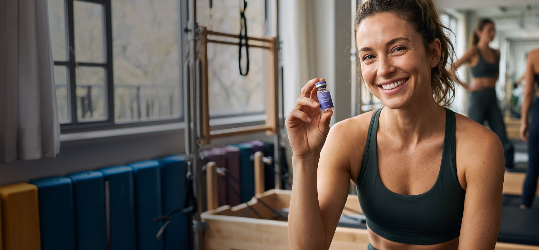 Smiling woman in dark green sports bra holding a small purple vial labeled 'Ivy Sermorelin Injection' in a gym with exercise equipment and mirrored walls.