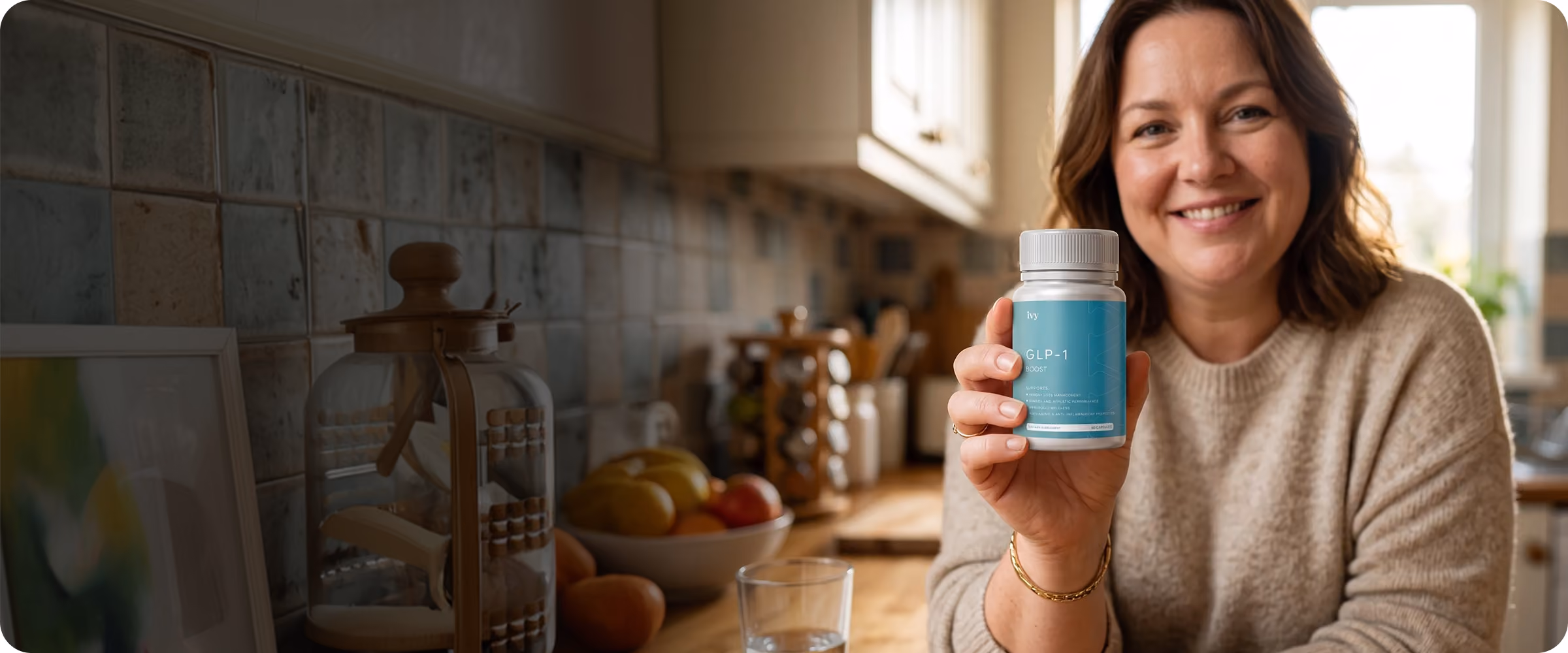 Smiling woman in a beige sweater holding a blue bottle labeled GLP-1 in a kitchen with fruit and glass on the counter.