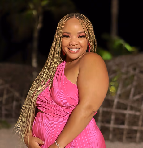 Smiling woman with long blonde braids wearing a bright pink sleeveless dress, posing outdoors at night with blurred dark background.