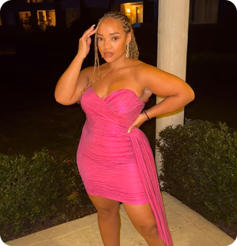 Woman with braided hair styled in cornrows wearing a strapless bright pink dress standing outside at night with one hand on her hip and the other near her temple, posing near a white pillar with greenery in the background.