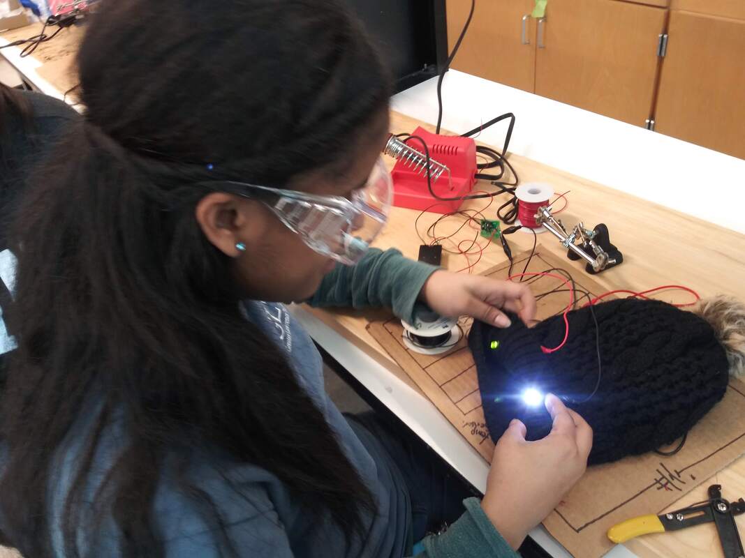 Person wearing safety goggles working on an electronic circuit connected to a black knitted hat with a glowing light.
