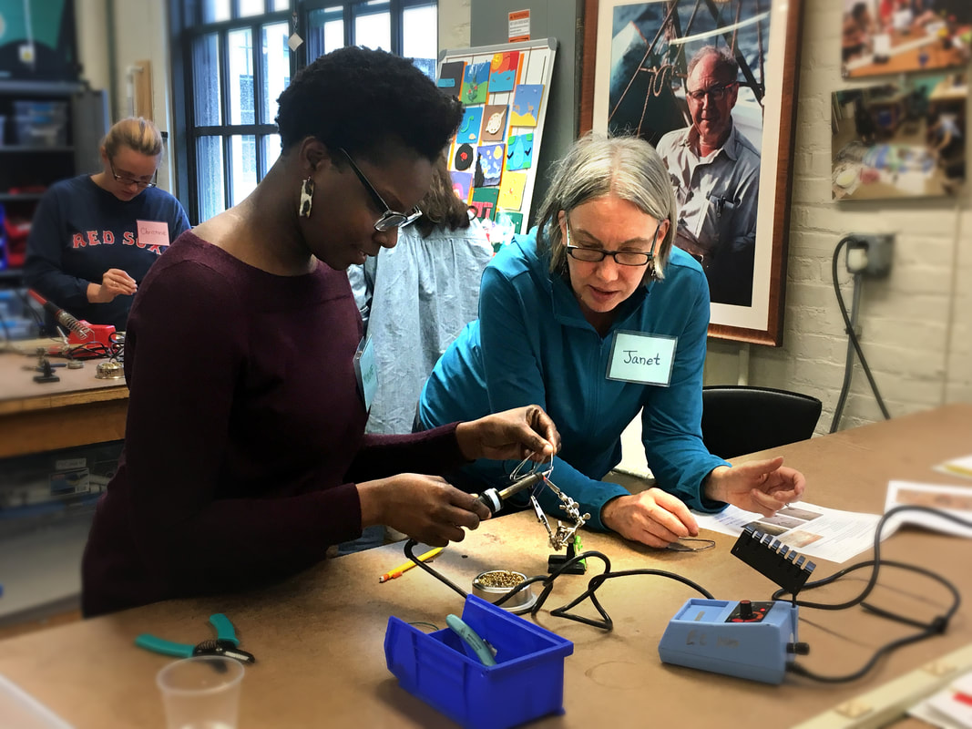 Two women working together on a small electronics project using a soldering iron at a workshop table.
