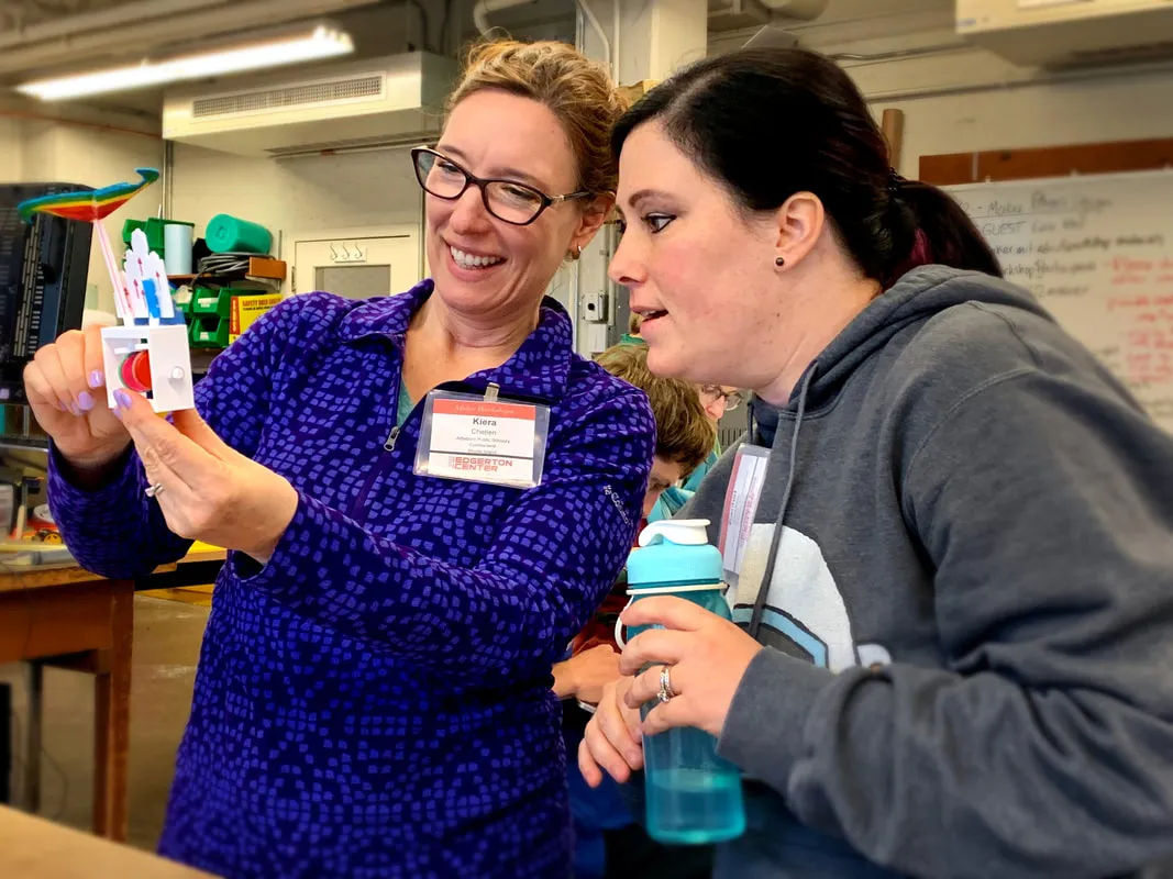 Two women in a workshop, one holding and showing a small colorful model to the other who is holding a blue water bottle.