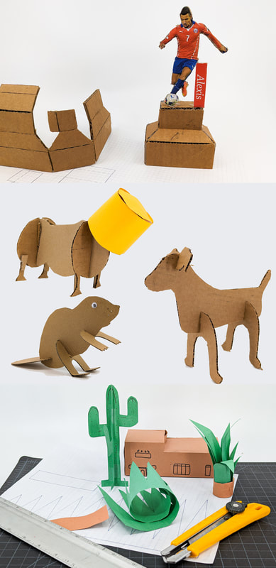 Collection of cardboard paper crafts including a soccer player on a pedestal, animals with a yellow cup on one, and a desert scene with cactus and house near cutting tools on a white surface.
