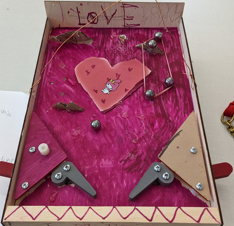 Homemade pinball machine with a pink heart and cartoon character at the center, decorated with purple color and metal balls.