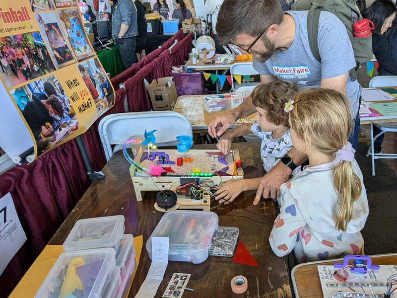 Man and two young girls interact with an illuminated electronic pinball-style game at a Maker Faire event.