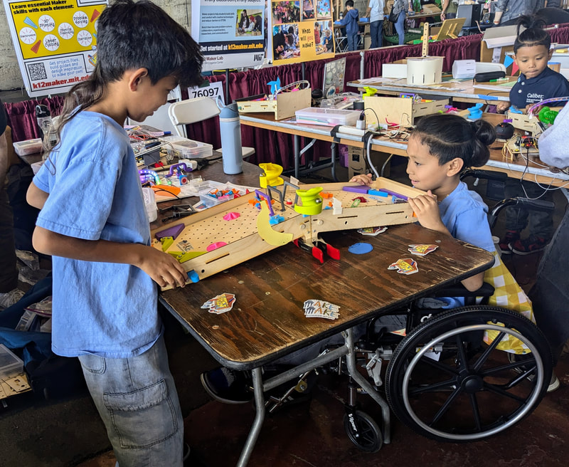 Two children engage with a wooden marble run game at a table, one child in a wheelchair, at an educational exhibit with posters and tables in the background.