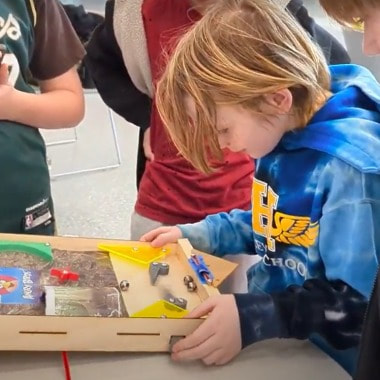 Young boy with blonde hair in a blue hoodie playing with a wooden pinball machine while others watch.