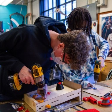 Two young people using cordless drills to work on a woodworking project indoors.