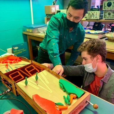 Two people wearing masks playing a tabletop pinball game indoors.