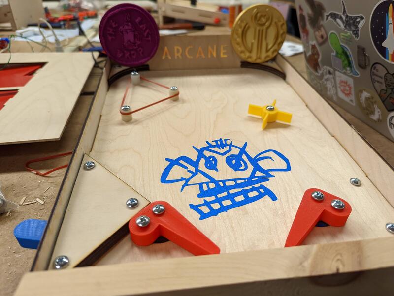 Wooden pinball game with orange flippers, yellow spinner, and blue doodle of a face in the center, labeled 'ARCANE' at the back.