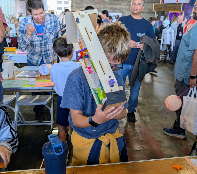 A boy examines a wooden mechanical model with colorful gears while people and tables are in the background at a busy indoor event.