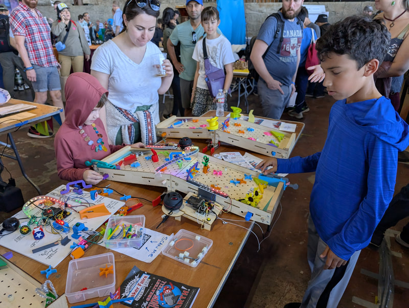Children and adults interacting with colorful, electronic marble maze games on tables at a crowded indoor event.