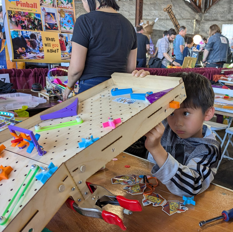 Young boy intently playing with a homemade wooden pinball board at a craft fair or workshop.