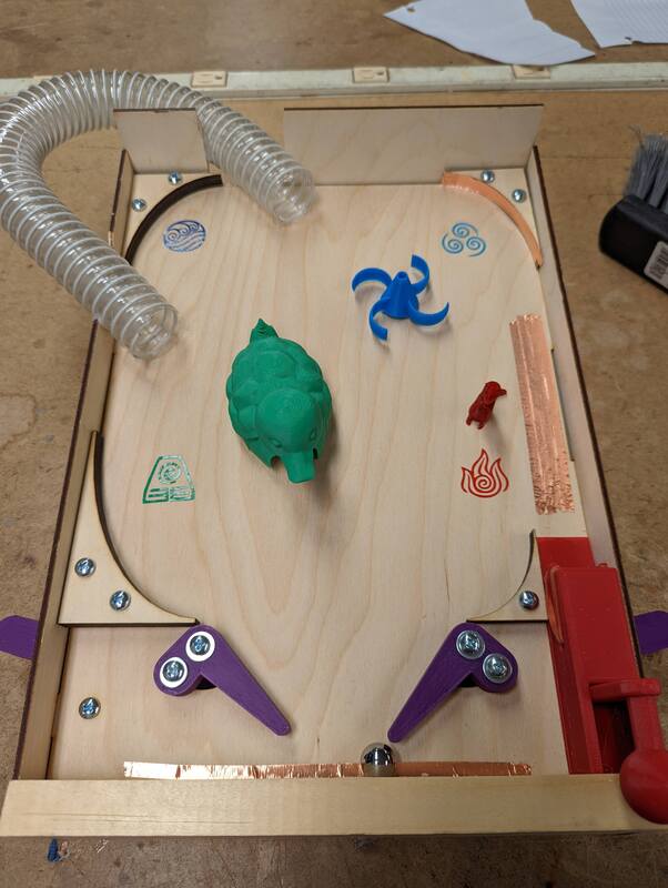 Wooden pinball machine with purple flippers, a green dinosaur figure, a blue propeller, a red toy dog, plastic tubing, and elemental symbols for water, air, earth, and fire.