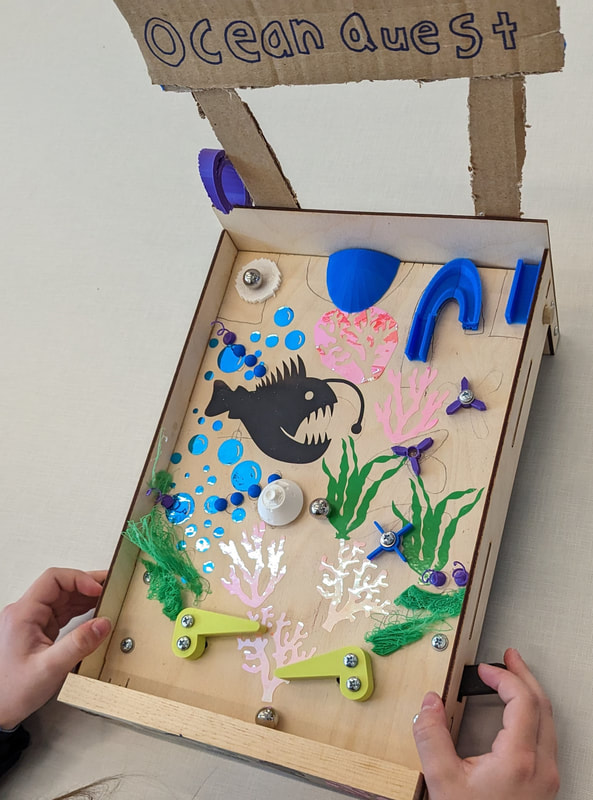 Hands holding a wooden pinball game decorated with ocean-themed elements including a black anglerfish silhouette and colorful coral designs.