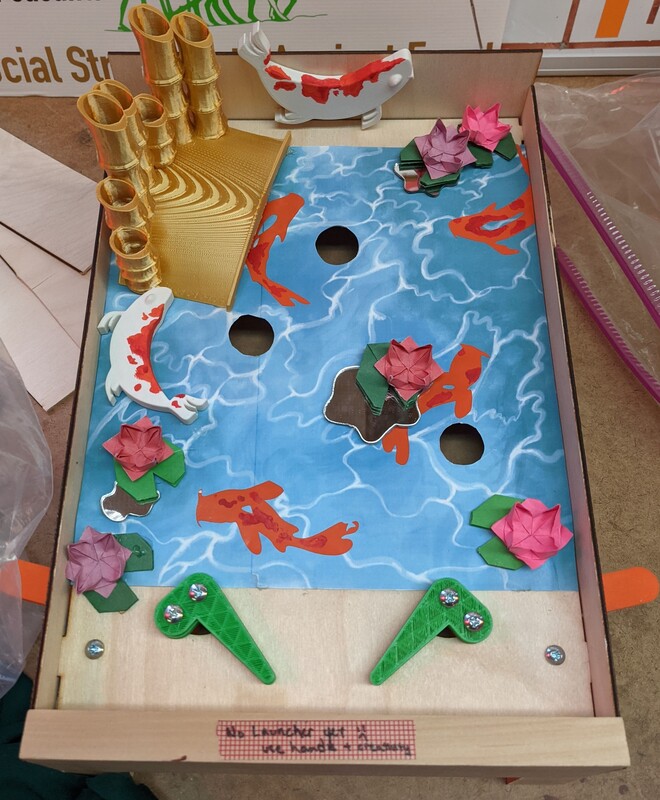 A tabletop pinball game with a blue water-themed surface featuring orange koi fish, red and purple lotus flowers, golden bamboo structures, and green flippers at the bottom.