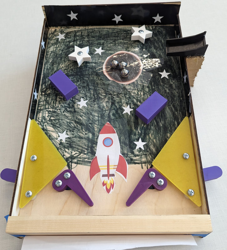 Homemade pinball game with a space theme featuring a rocket, stars, yellow flippers, purple bumpers, and steel ball bearings inside a cardboard box.