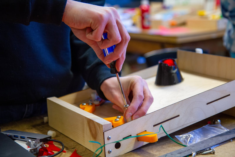 Person using a screwdriver to assemble a wooden box with orange components and wires on a cluttered workbench.