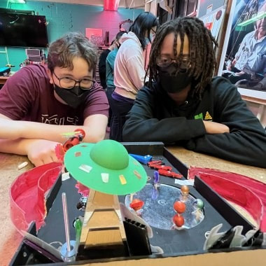 Two people wearing black face masks sitting at a table playing a tabletop game with a green flying saucer piece.