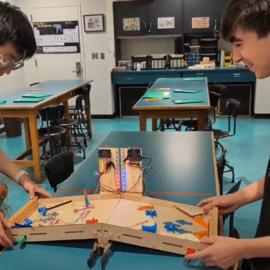 Two young men working together on a tabletop robotics project with LEDs on a wooden circuit board in a classroom.