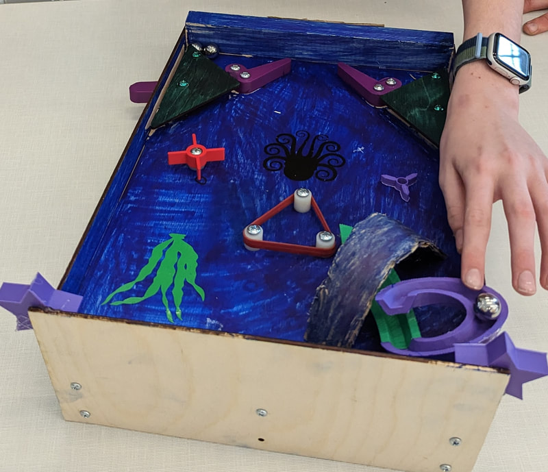 A hand near a handmade wooden pinball machine with blue painted inside, green and purple details, and metal balls.