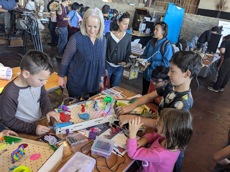 A group of children and adults gathered around a table engaged in a hands-on STEM activity with electronic components and colorful toys.