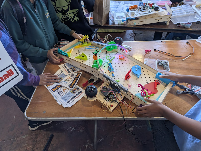 Three people playing with a colorful pinball machine prototype on a wooden table with electronics and flyers nearby.