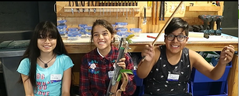 Three smiling children sitting in front of a workshop table with tools and plastic containers, one holding a handcrafted string instrument.