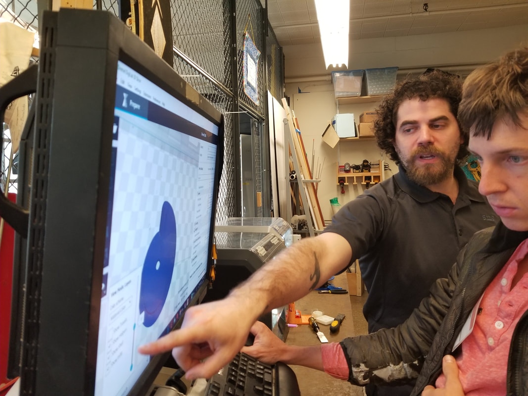 A man with curly hair and a beard points at a computer screen showing 3D modeling software as a young man watches intently in a workshop.