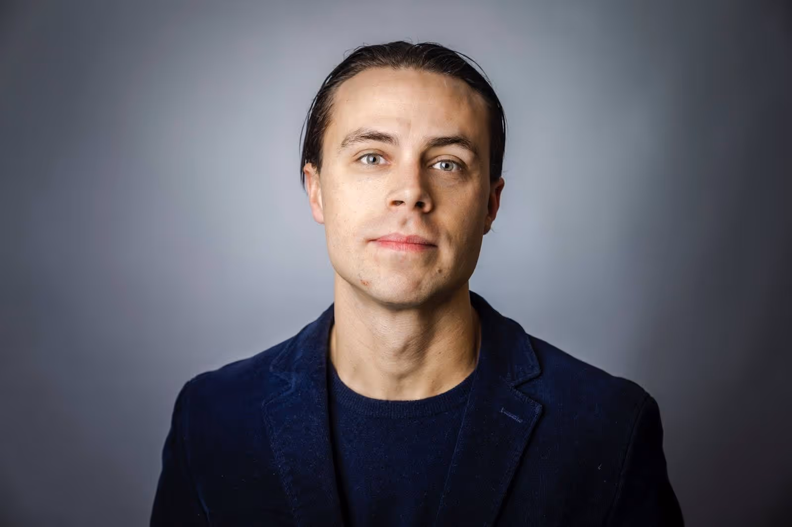 Portrait of a man with slicked-back hair wearing a dark blazer and navy shirt against a gray background.