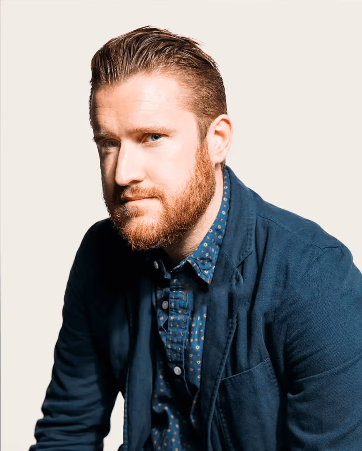 Man with red hair and beard wearing a navy blazer over a patterned shirt, looking forward.