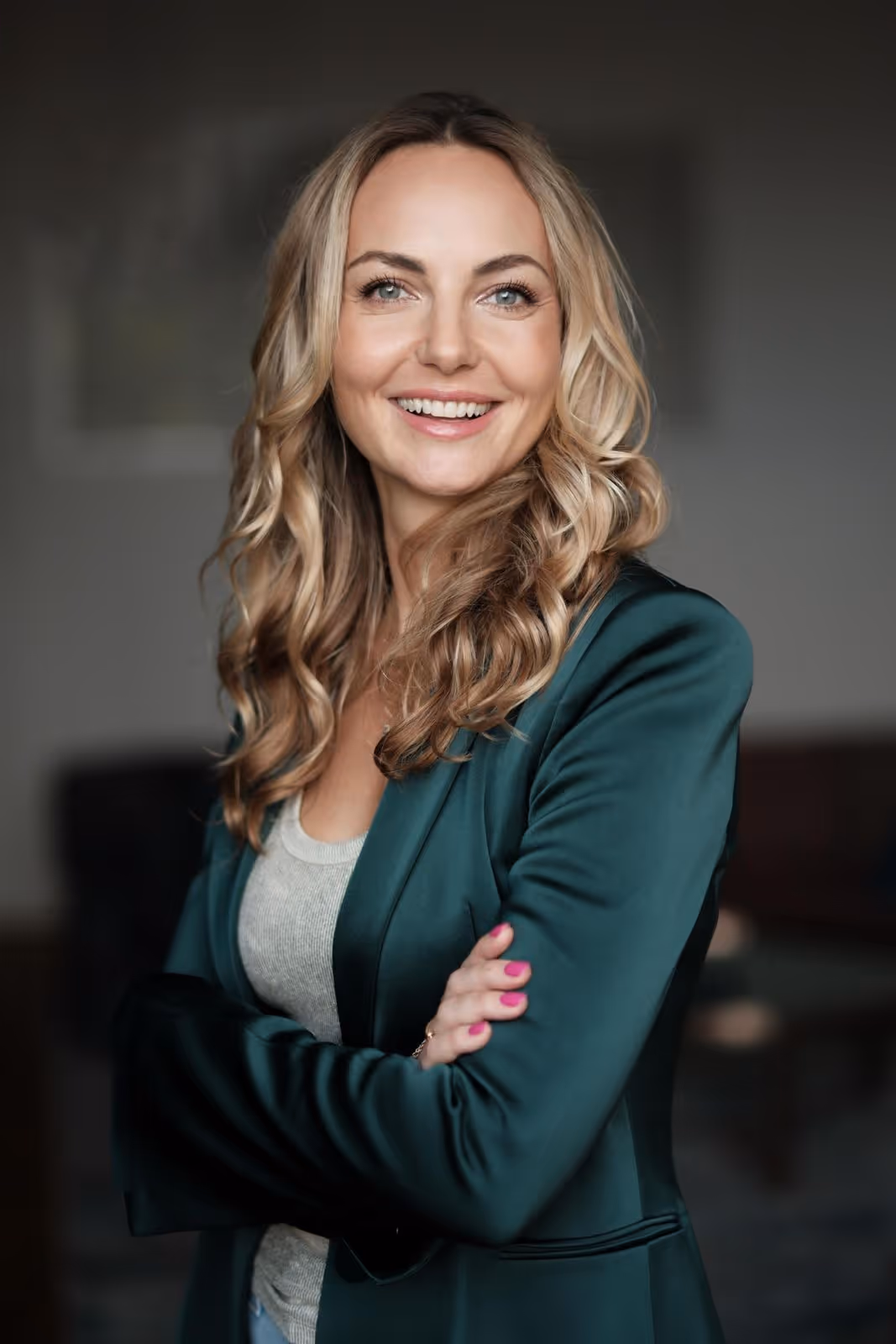 Smiling woman with blonde curly hair wearing a green blazer and crossed arms in a blurred indoor setting.