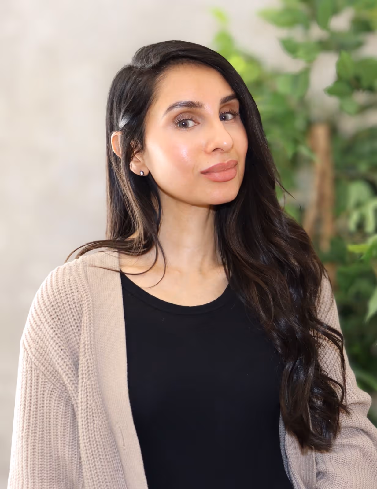 Woman with long dark hair wearing a beige sweater over a black top, posing indoors with greenery behind.