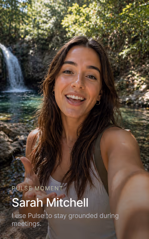 Smiling woman with long brown hair outdoors near waterfall and rocks, taking a selfie.