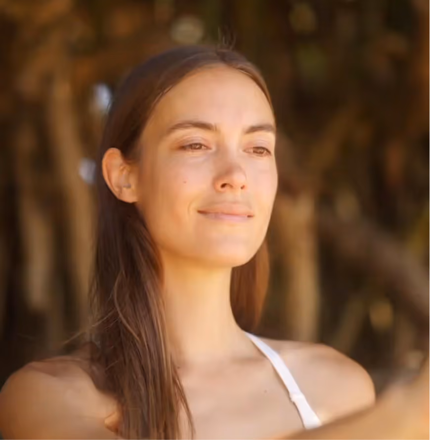 Woman with long brown hair looks content with a blurred natural background behind her.