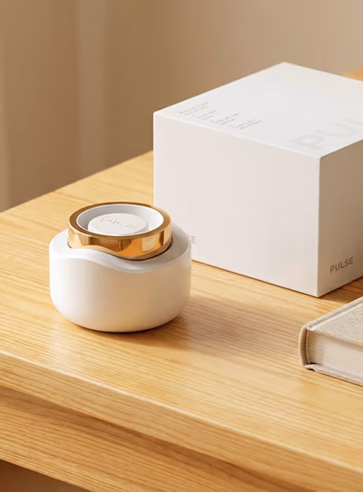 White and gold Pulse device on wooden table near white box and closed beige book.