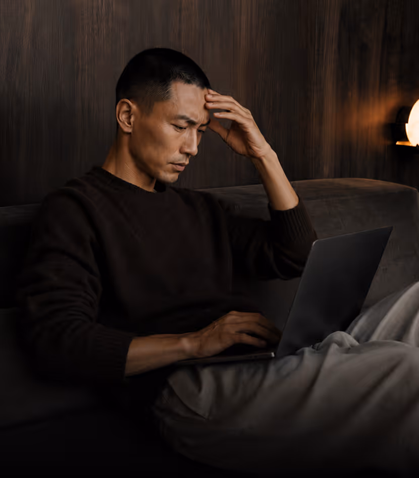Man sitting on a couch using a laptop with a concerned expression, hand on forehead.