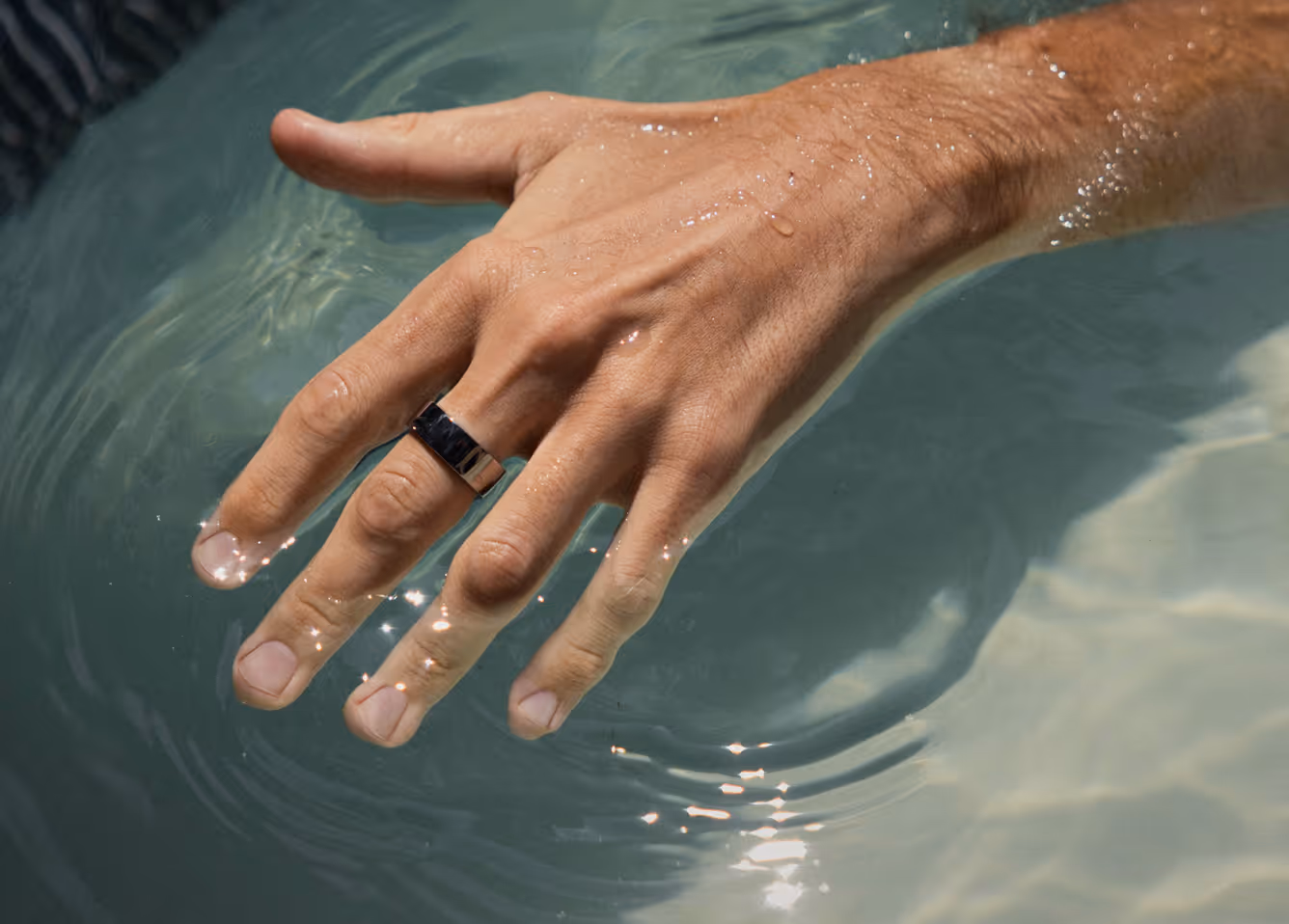 Close-up of a wet hand with a black ring dipping in water creating ripples and light reflections.