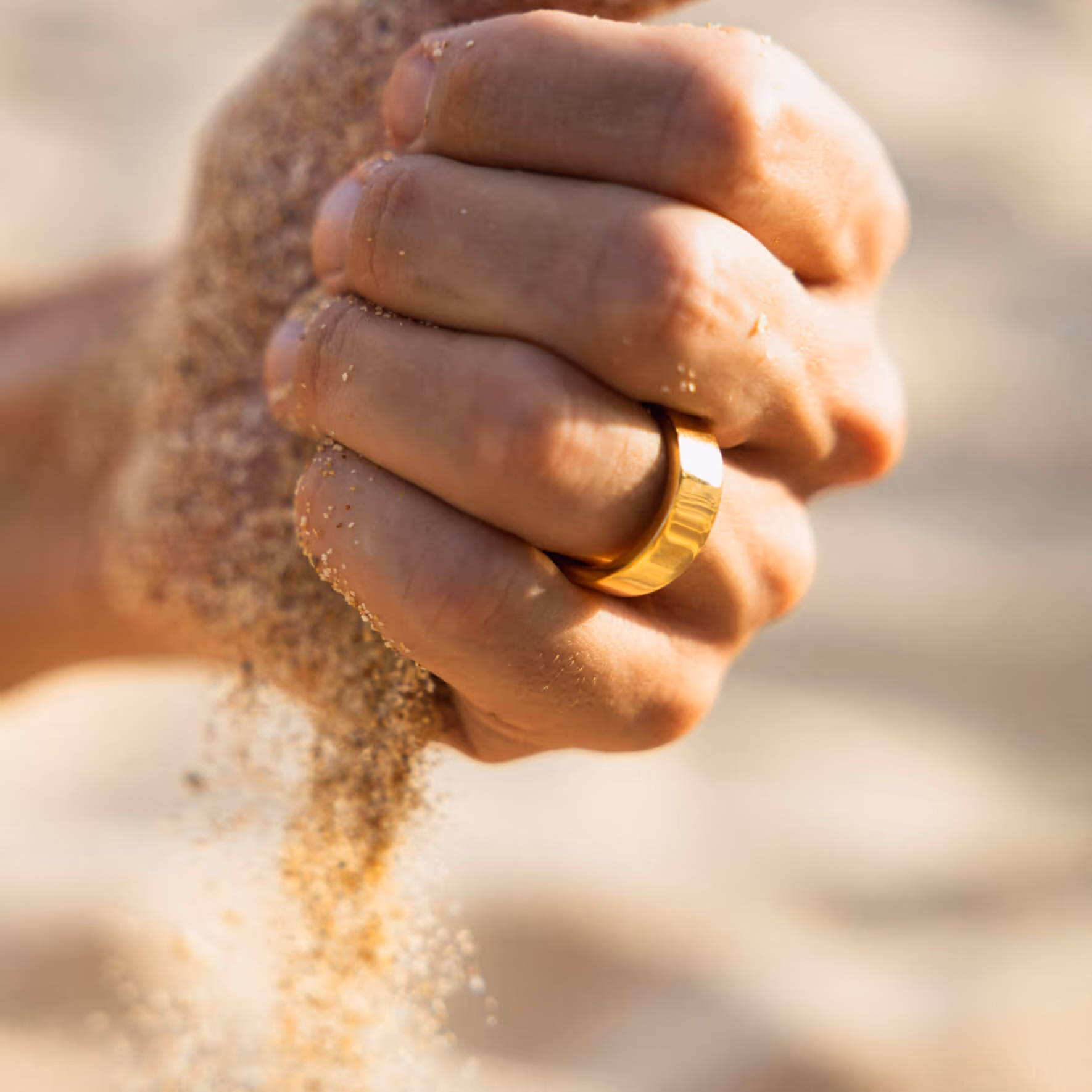 Hand with gold ring letting sand fall between fingers on a beach.