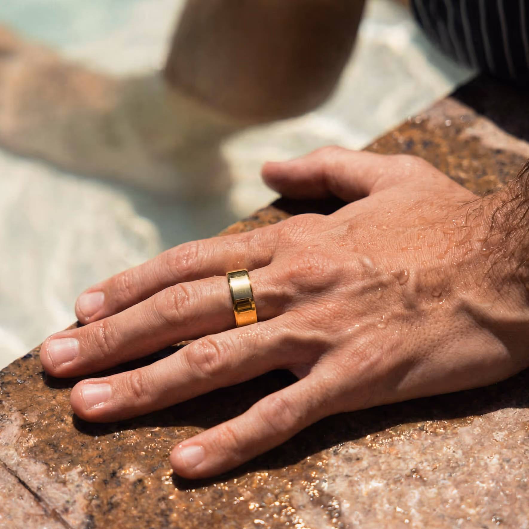 Close-up of a wet hand wearing a wide gold ring resting on a textured stone surface near water.