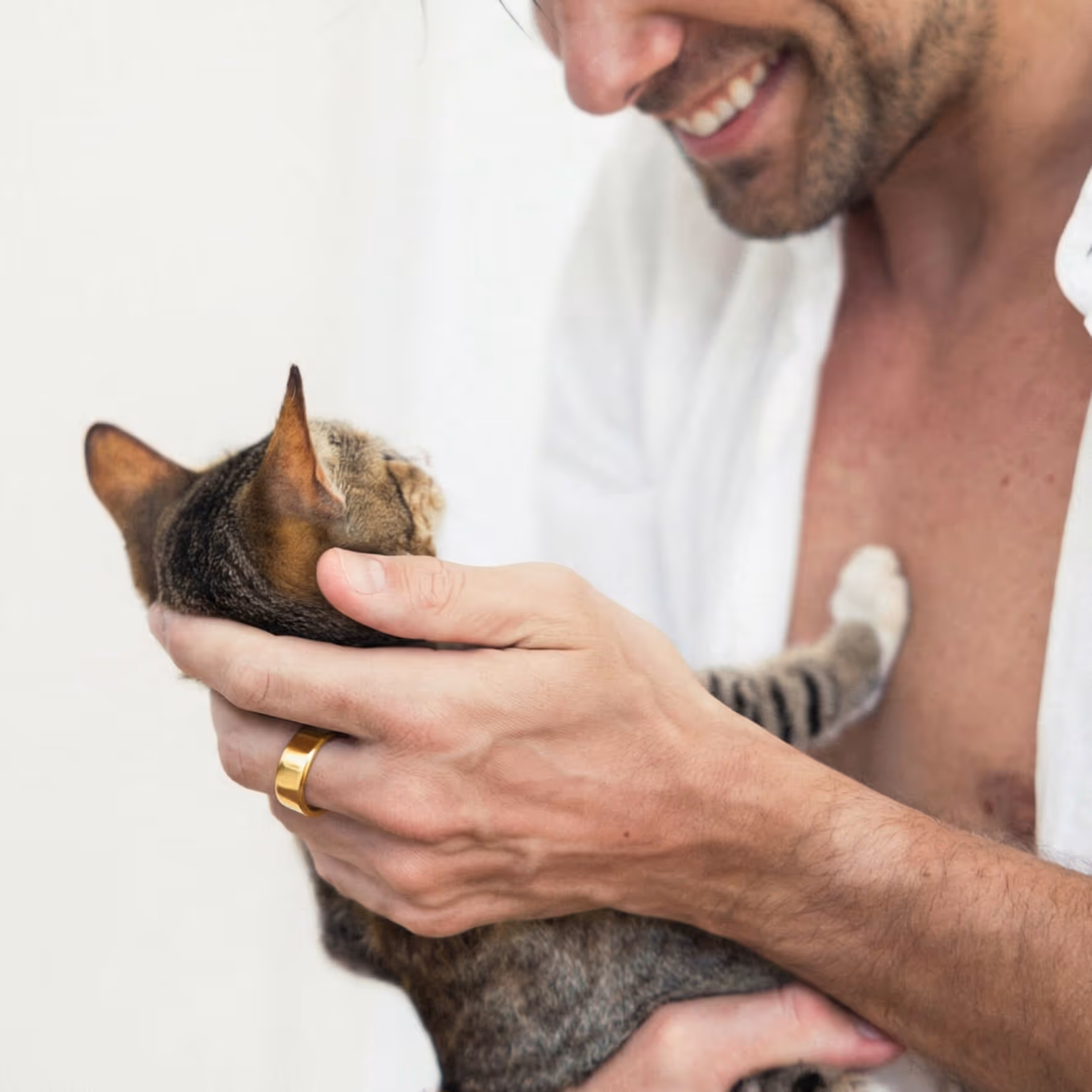 Man wearing a gold ring holding and smiling at a tabby cat against a white background.