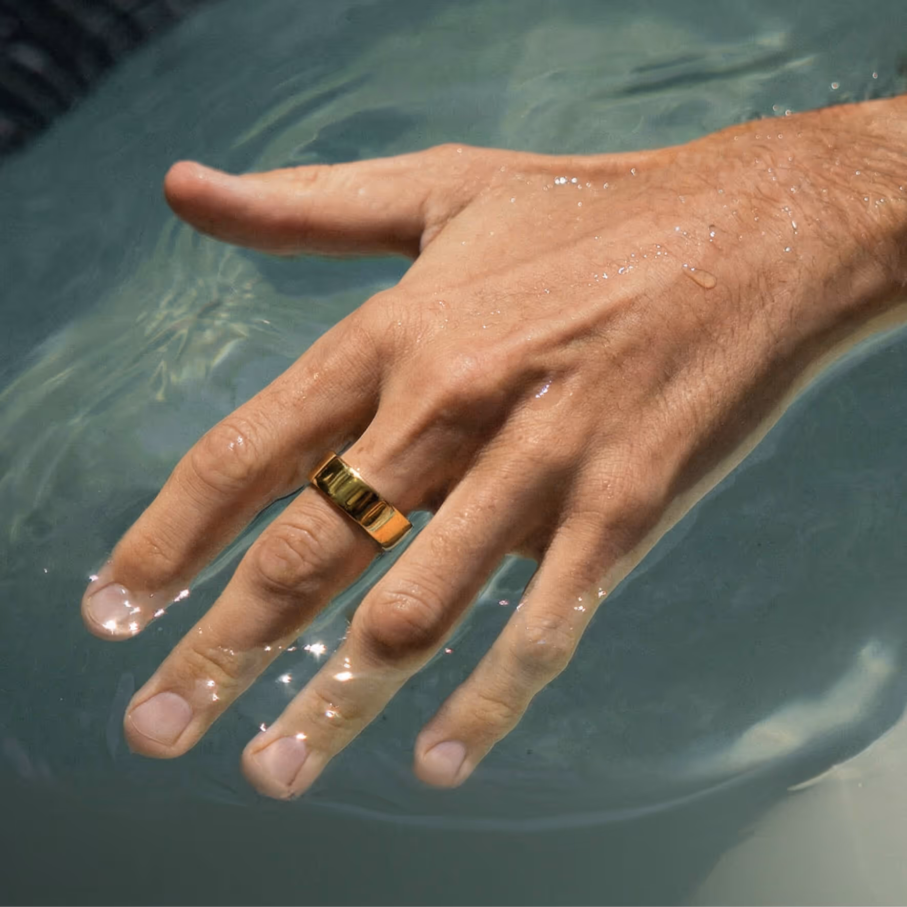 Male hand partially submerged in water wearing a wide gold ring on the ring finger.
