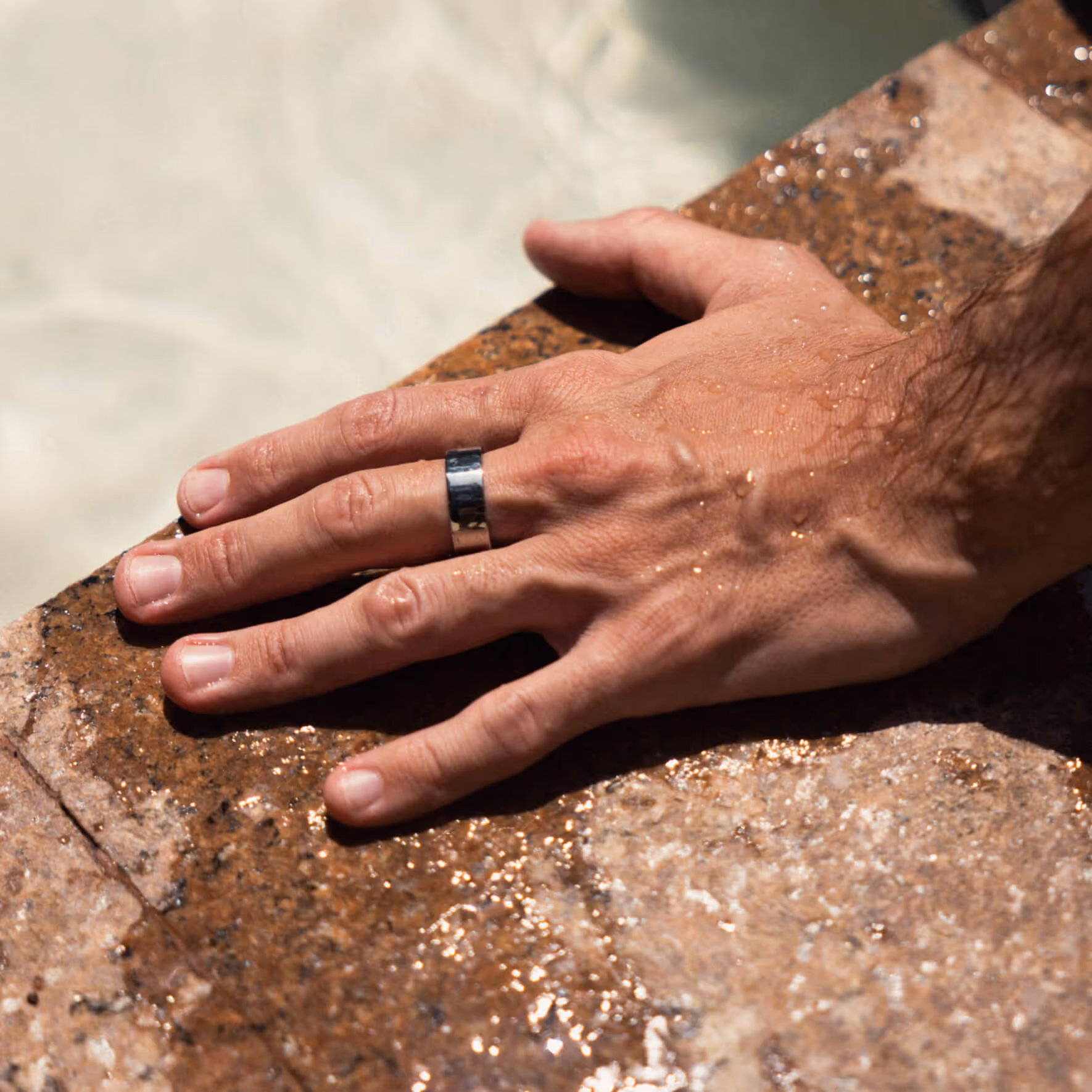 Male hand with a silver ring resting on a wet stone surface near water.