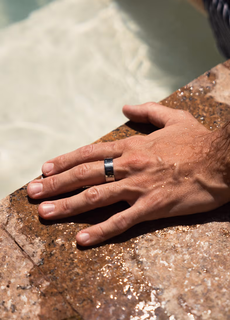 Close-up of a wet hand with a silver ring resting on a wet stone surface near water.