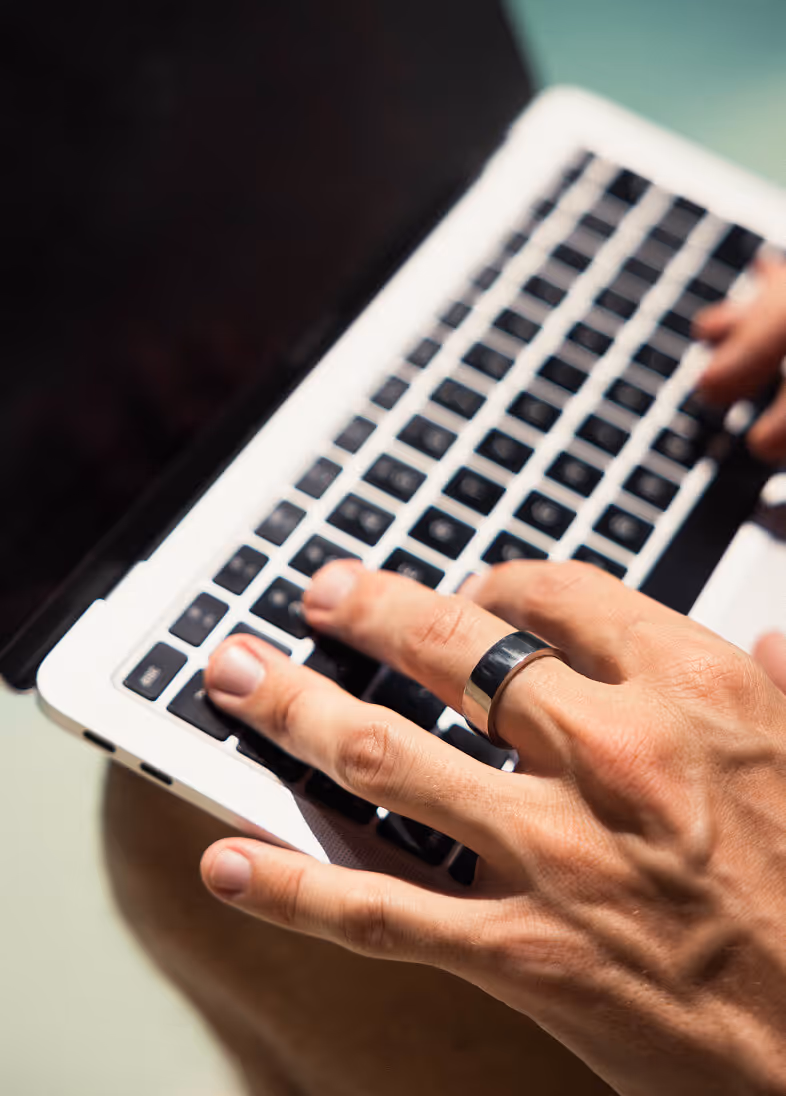 Right hand wearing a shiny silver ring typing on a laptop keyboard with black keys.