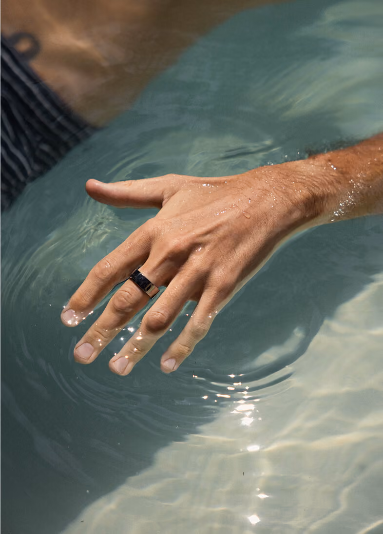 Male hand with a silver ring dipping fingers into clear water, creating ripples and reflections.