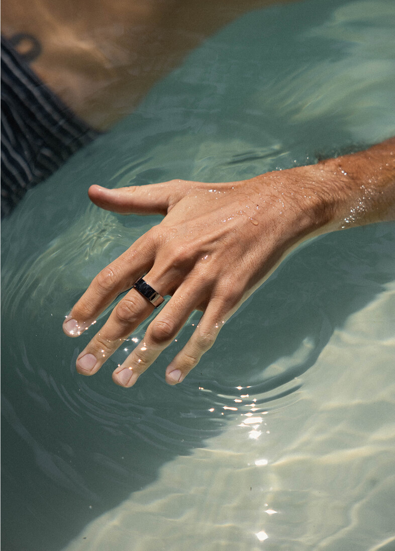 Male hand with a silver ring dipping fingers into clear water, creating ripples and reflections.